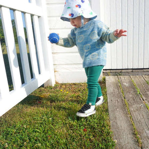 toddler walking with blue tennis ball wearing smiley jacquard sweater, printed bucket hat and green organic cotton leggings