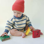 Load image into Gallery viewer, baby wearing an organic cotton ivory and indigo blue chunky striped sweater and a red alpaca/wool knit beanie sitting on the floor and playing with bright colorful board books
