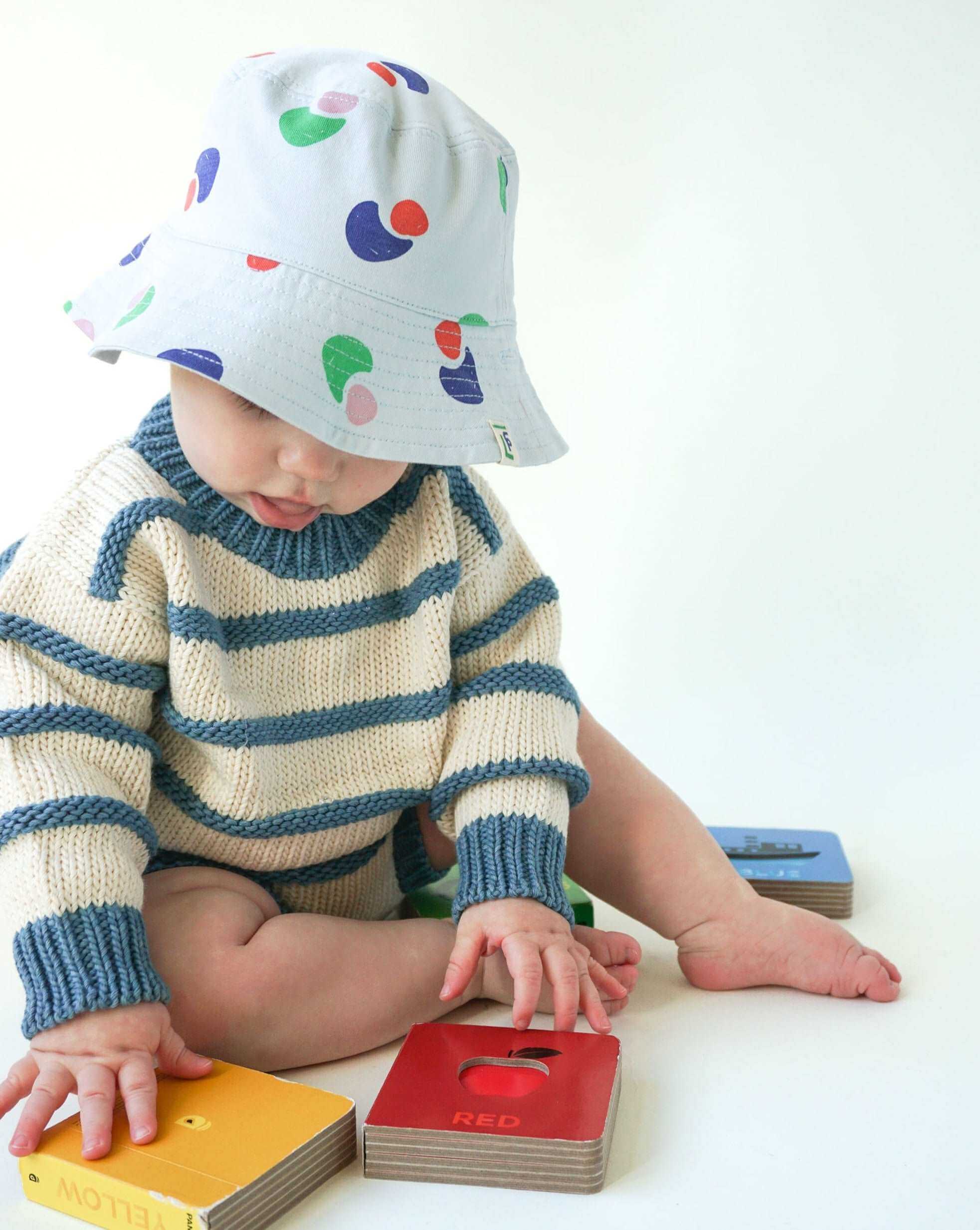 baby wearing an organic cotton ivory and indigo blue chunky striped sweater and a bright printed baby blue bucket hat sitting on the floor and playing with bright colorful board books
