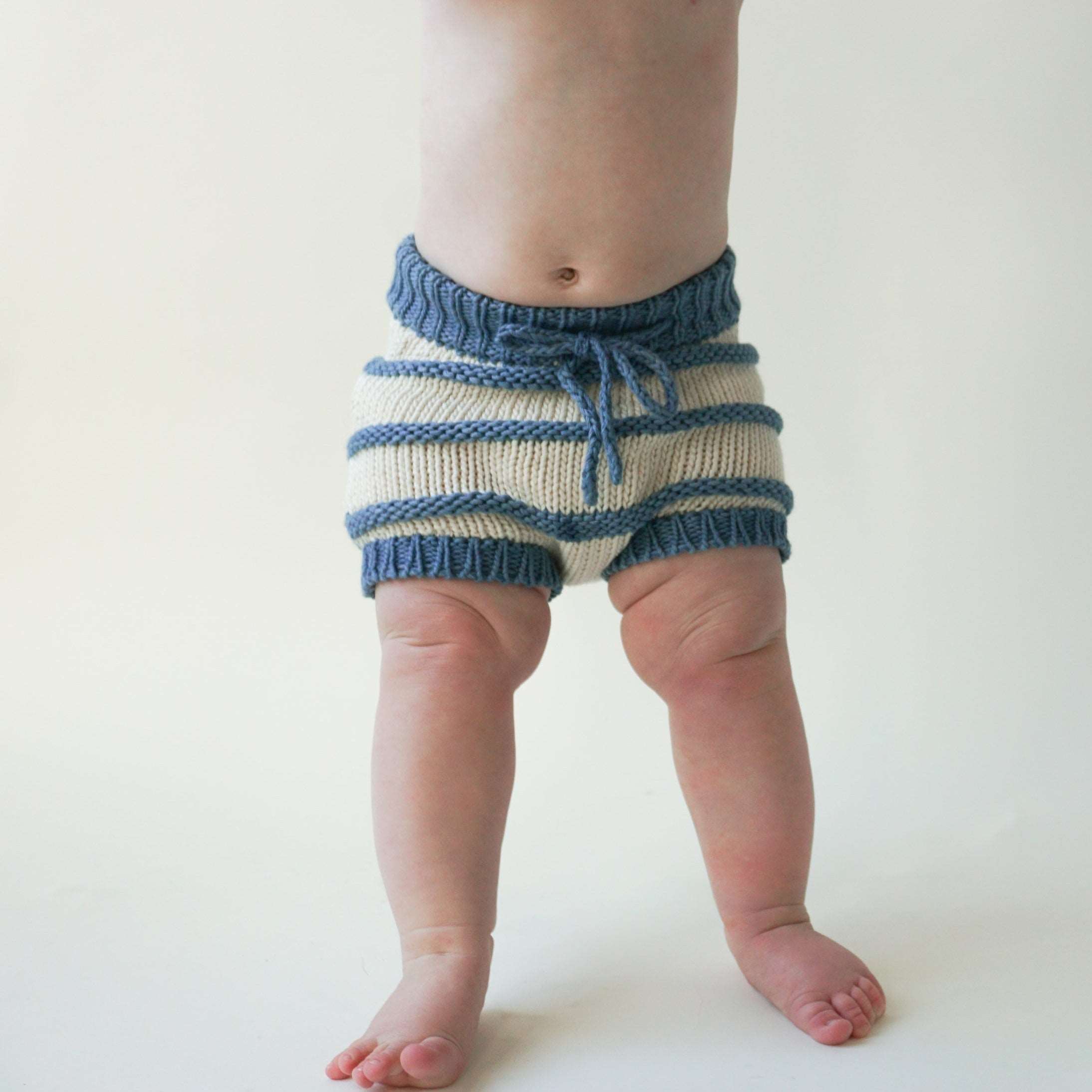 View of a baby, chest and down standing and wearing only an ivory and indigo striped organic cotton chunky knit bloomer with indigo blue waistband and drawstring.
