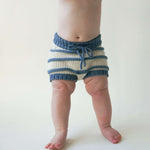 Load image into Gallery viewer, View of a baby, chest and down standing and wearing only an ivory and indigo striped organic cotton chunky knit bloomer with indigo blue waistband and drawstring.
