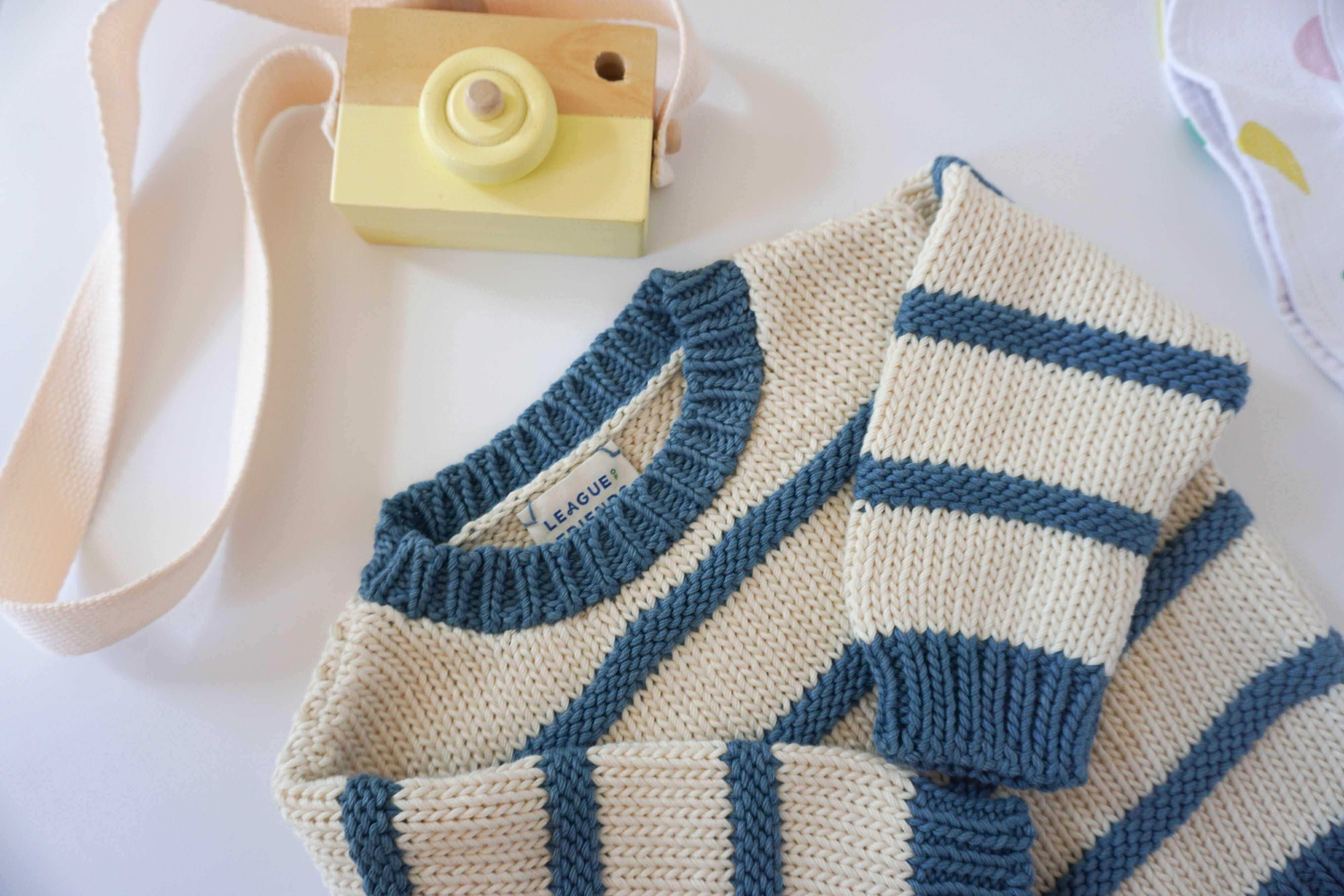 ivory and indigo blue organic cotton chunky striped sweater laying on background with sleeves folded and a yellow wooden toy camera sitting next to the sweater on the background