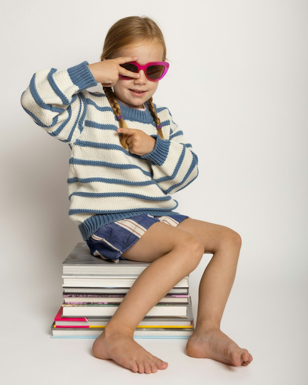 A young girl with braided hair sits on a stack of books against a plain light gray studio background. She is wearing a blue and white striped knit sweater, denim shorts, and bright pink heart-shaped sunglasses, holding one lens up to her eye.