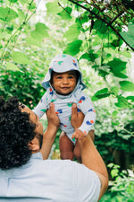 Load image into Gallery viewer, Back view of man with curly hair holding smiling baby wearing a bright colorful printed baby blue wrap onesie bodysuit and bucket hat with green bushes and trees in the background.
