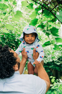 Back view of man with curly hair holding smiling baby wearing a bright colorful printed baby blue wrap onesie bodysuit and bucket hat with green bushes and trees in the background.