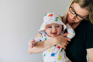 Mom wearing black t-shirt and glasses smiling while holding happy baby wearing primary colored printed organic cotton bucket hat and wrap bodysuit.