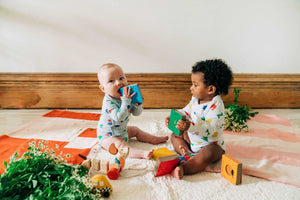 two babies wearing colorful organic cotton bodysuits playing with colorful books and sitting on blankets