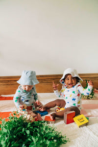two happy babies wearing printed organic cotton wrap bodysuits and solid white and baby blue bucket hats sitting on striped blankets and playing with colorful board books and a wooden dog.