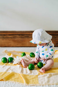 baby sitting and playing with limes on a yellow striped blanket wears a primary color printed organic cotton wrap bodysuit and a solid white bucket hat that has a printed reverse side.