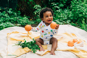 Smiling baby wearing a colorful organic cotton long sleeve onesie bodysuit sitting on a yellow blanket holding two oranges with green plants in the background