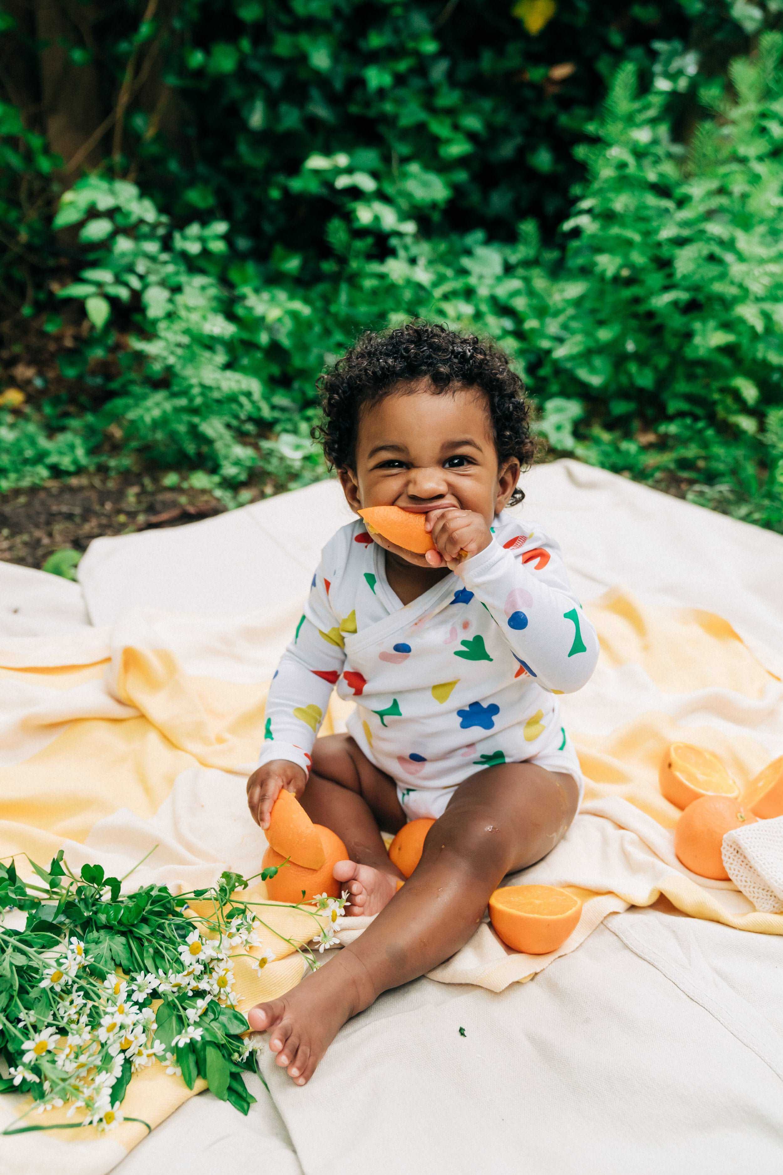 cute baby in rainbow colored organic cotton wrap bodysuit onesie eating orange slice and sitting on yellow blanket