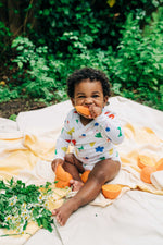 Load image into Gallery viewer, cute baby in rainbow colored organic cotton wrap bodysuit onesie eating orange slice and sitting on yellow blanket
