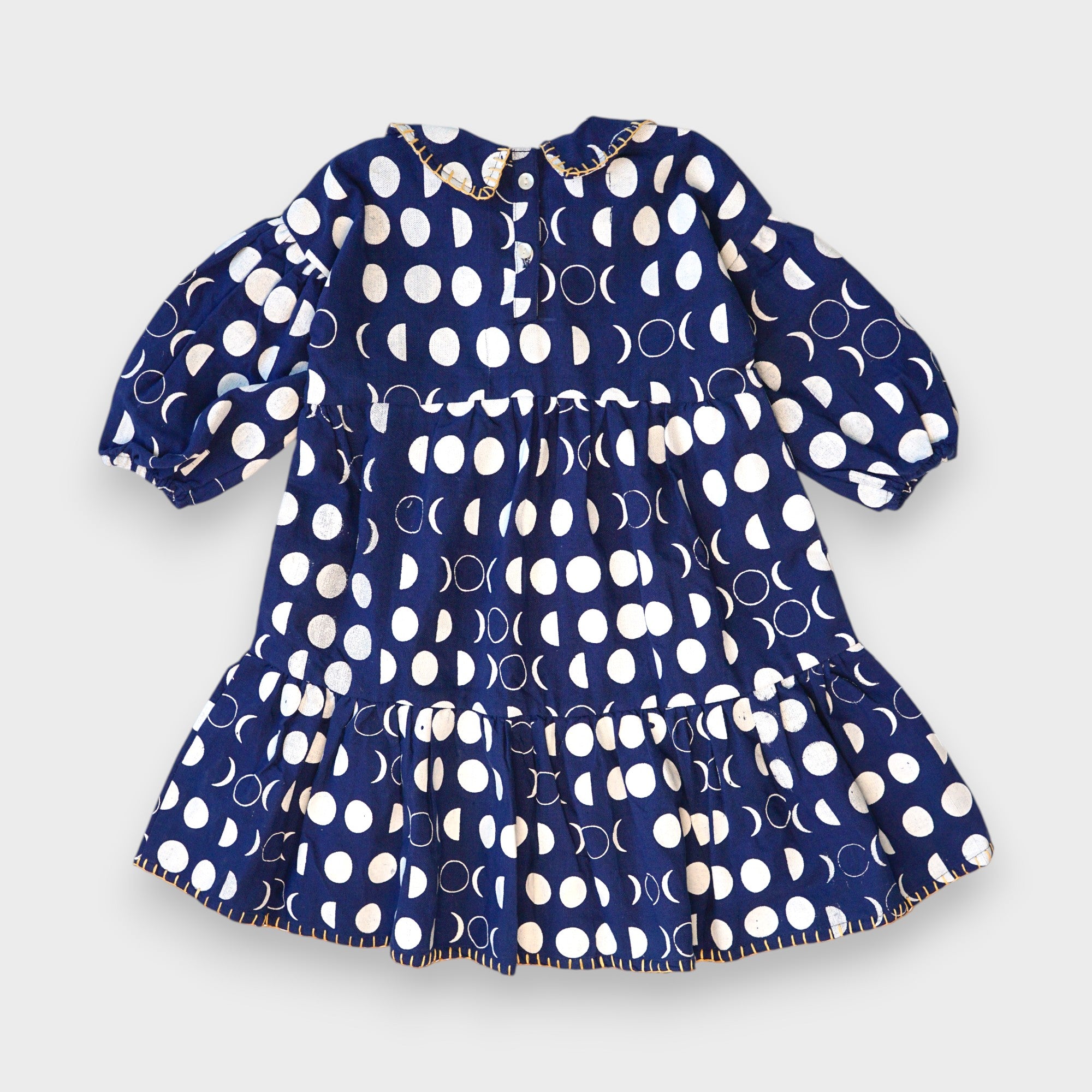 Back of navy Moon Phases tiered dress with cream moon print, back buttons, puff sleeves and yellow-stitched hem.