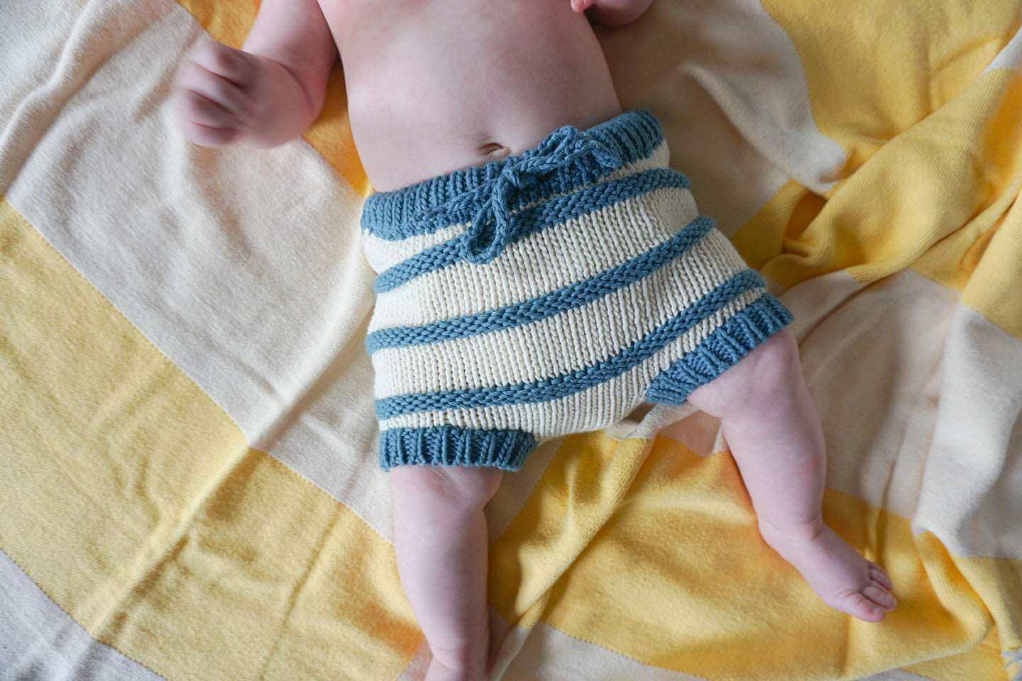 Front view of a baby laying on a yellow and cream striped blanket, wearing only an ivory and indigo blue striped organic cotton chunky knit bloomer short.