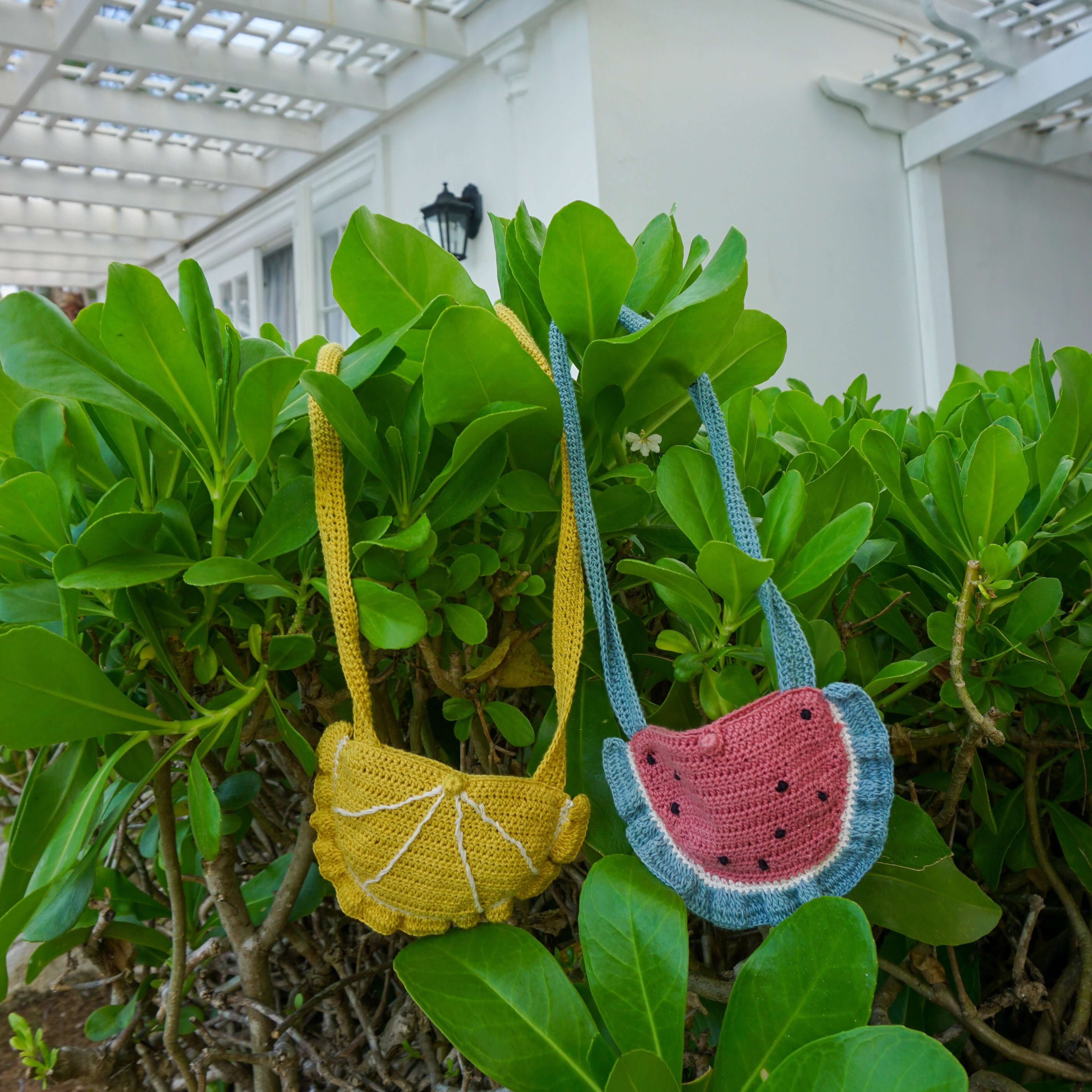 two crochet watermelon and lemon purses hanging on a green bush