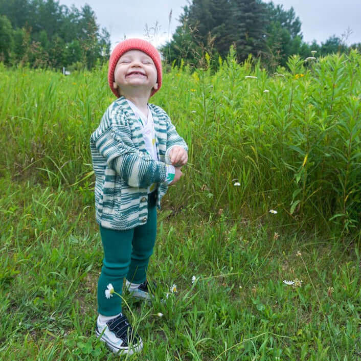 Smiling baby wearing melange cable knit v-neck cardigan, red wool and baby alpaca beanie and organic cotton leggings