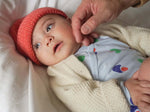 Load image into Gallery viewer, Baby wearing red knit beanie hat and organic cotton wrap onesie and ivory wool/alpaca cardigan
