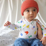 Load image into Gallery viewer, baby sitting wearing a red beanie and a rainbow colored wrap onesie
