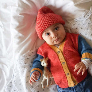 Baby laying down wearing red wool/alpaca beanie hat and red, yellow, blue colorblocked cardigan