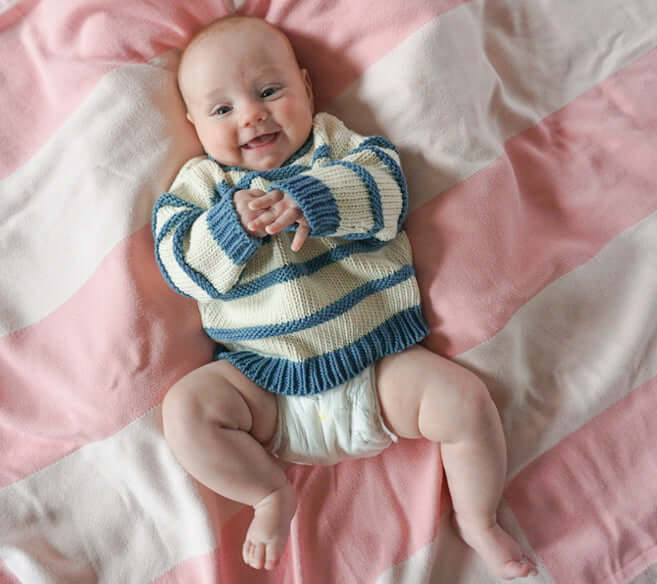smiling baby, laying on a pink striped blanket, wearing an organic cotton chunky ivory and indigo blue striped sweater and a diaper, clapping their hands together.